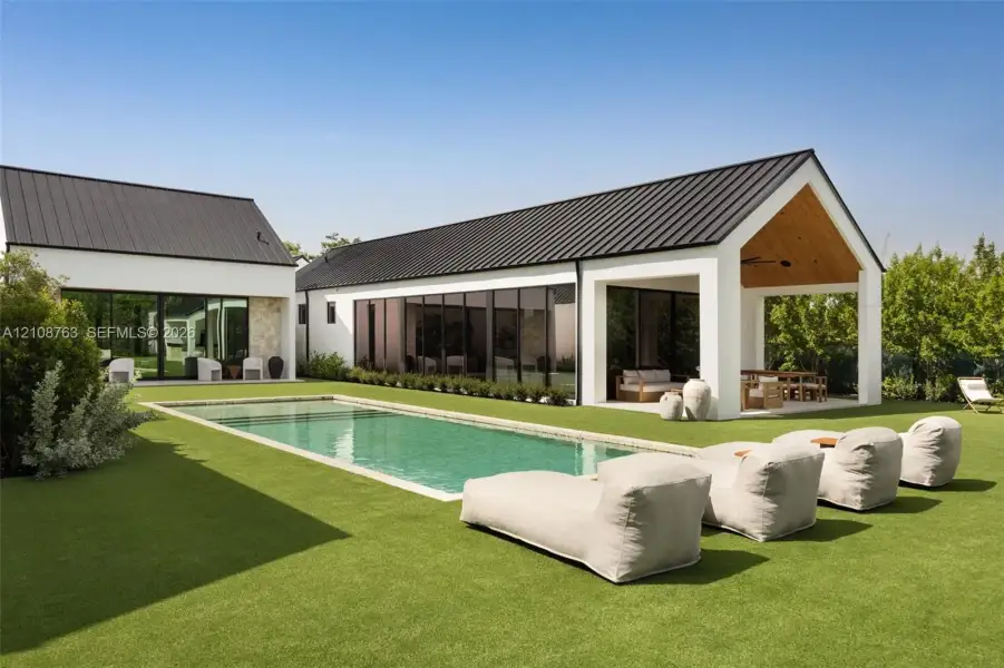 Rectangular in-ground swimming pool, metal roof, covered patio with wood-paneled ceiling, large-format glass wall panels, and expansive lawn