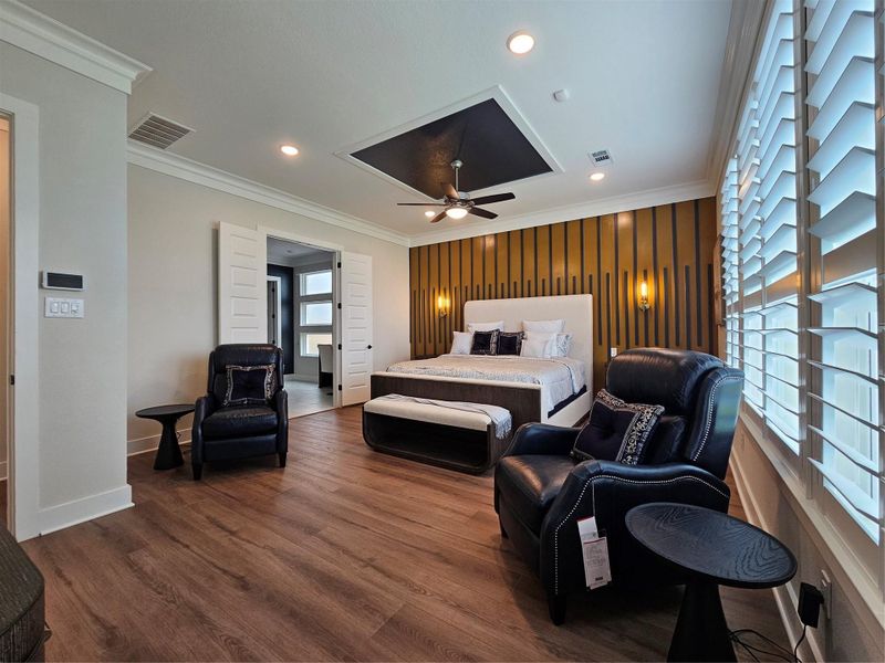 This spacious bedroom features elegant wood flooring, a modern accent wall behind the bed, and ample natural light from large windows with plantation shutters. It includes comfortable seating with two leather chairs and side tables, and there's a ceiling fan for added comfort. The room connects to an adjoining space through stylish double doors. This spacious bedroom features elegant wood flooring, a modern accent wall behind the bed, and ample natural light from large windows with plantation shutters. It includes comfortable seating with two leather chairs and side tables, and there's a ceiling fan for added comfort. The room connects to an adjoining space through stylish double doors.