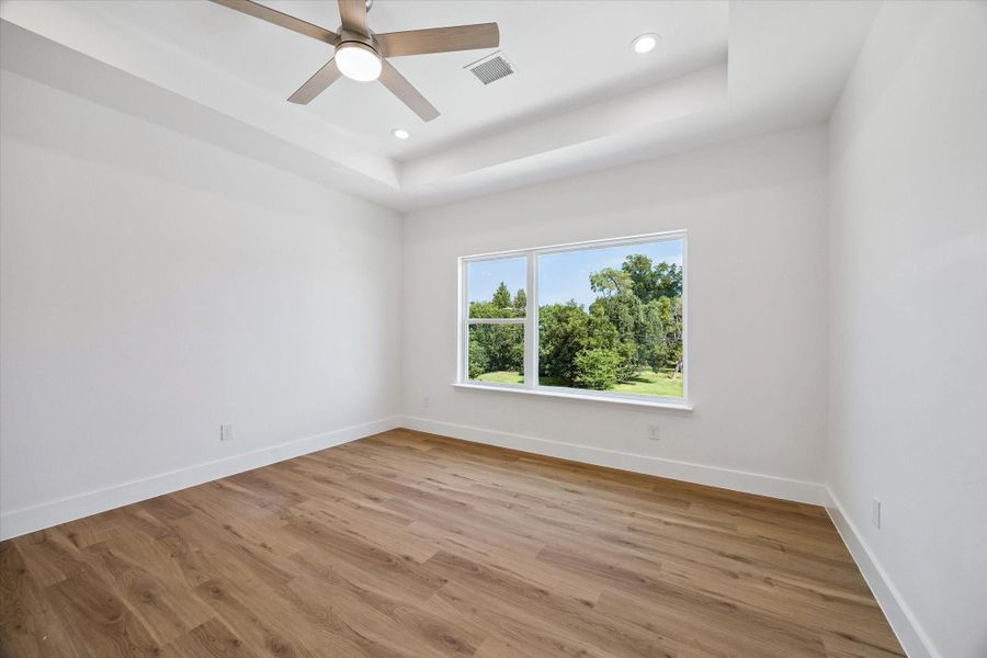 Another view of the bedroom highlights its generous proportions, elevated tray ceiling, and abundant natural light. The large window frames leafy outdoor views while warm wood-look flooring adds character and continuity. A contemporary ceiling fan and recessed lighting complete the clean, modern presentation. The open floor plan provides flexibility for substantial furniture, a sitting area, or additional storage pieces.