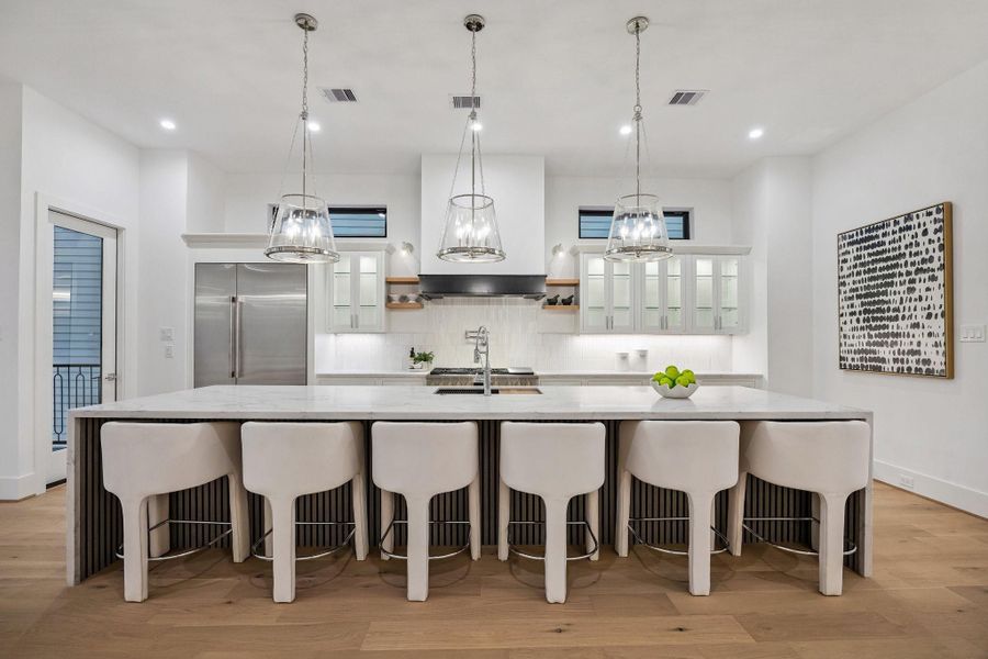 This stunning chef's kitchen showcases a dramatic Quartz waterfall island with ample seating, perfect for entertaining.  Crisp white inset cabinetry and glass front storage maintain visual lightness, while black accents and Polished Nickel pendant lighting introduce depth and structure.  Stainless Appliances, custom hood, Pot Filler, under-cabinet lighting, warm wood floors, and abundant natural light complete this elegant, modern space.