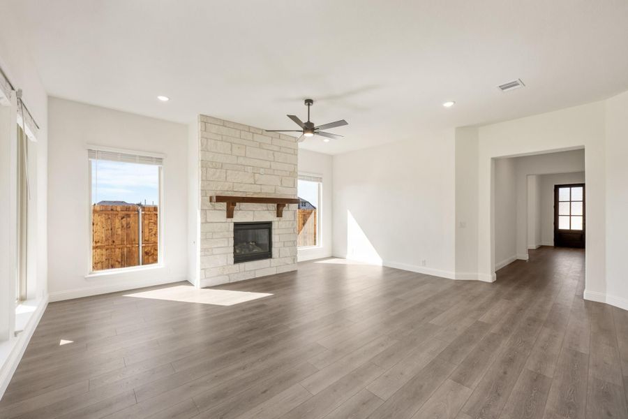 Open living room with stone fireplace, wood mantel, ceiling fan, and gray hardwood floors Open living room with stone fireplace, wood mantel, ceiling fan, and gray hardwood floors