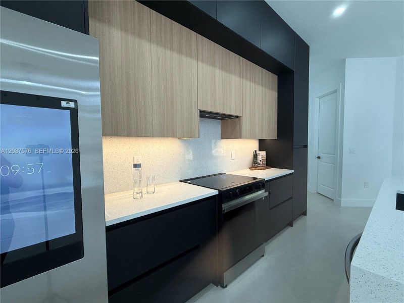 Contemporary kitchen featuring dark flat-panel cabinetry, light wood-finish upper cabinets, white speckled countertops, range and stainless steel appliances, range hood