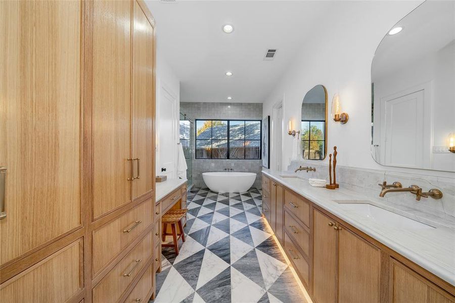 Spacious bathroom featuring a double vanity with stone countertops and undermount sinks, wood-finish cabinetry, geometric tile flooring, and a freestanding soaking tub