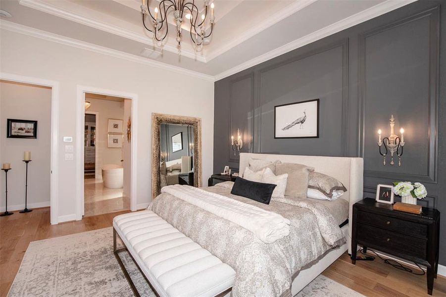 This photo showcases a stylish bedroom with a sophisticated design. It features a plush bed with elegant bedding, a dark accent wall with sconce lighting, and a decorative chandelier. The room has hardwood flooring and an adjoining bathroom, providing a luxurious and comfortable living space.