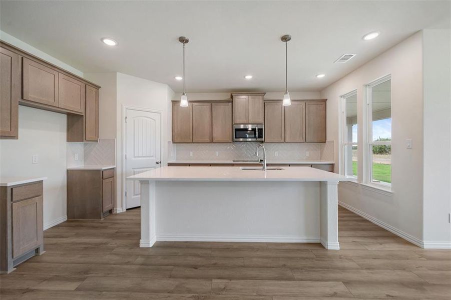 Kitchen with pendant lighting, backsplash, stainless steel microwave, a center island with sink, and light wood-style floors