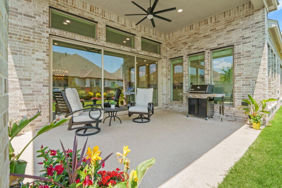 This photo showcases a spacious, covered patio with room for comfortable seating, a ceiling fan, and a barbecue grill, ideal for outdoor entertaining. Surrounded by well-maintained landscaping, the area offers a tranquil setting with large windows providing a view into the interior.