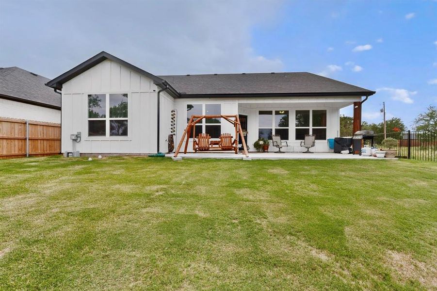 Expansive backyard featuring a covered concrete patio, wood-finish support column, white board and batten siding, dark-framed windows, and a dark shingle roof