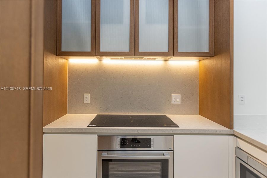 Kitchen area featuring matte white base cabinetry, integrated stainless steel oven, cooktop, solid-surface countertop, and wood-finish upper cabinets with fluted glass panel inserts