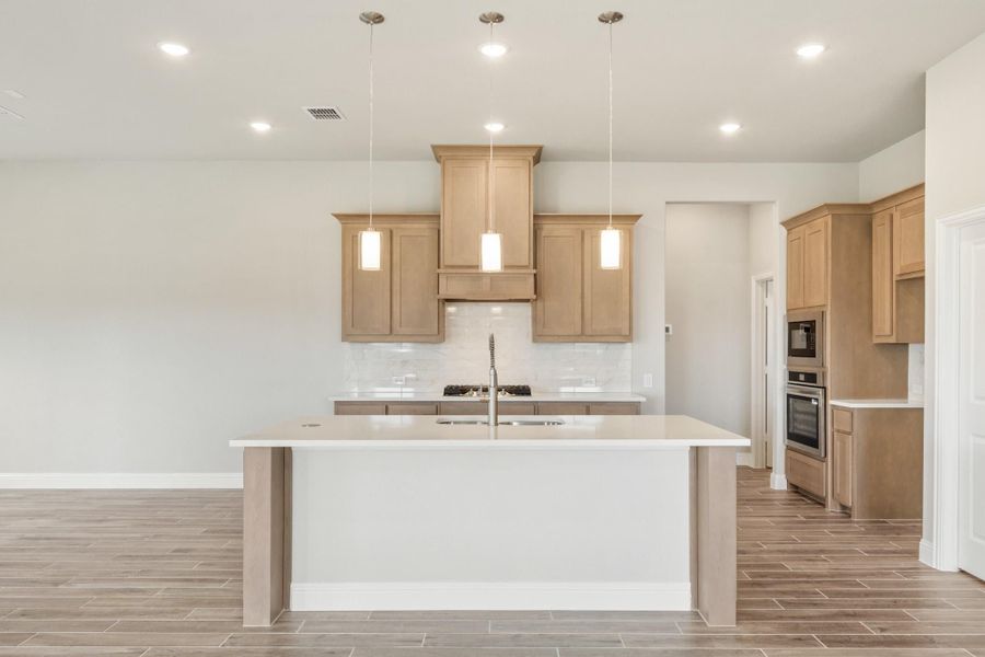 Kitchen with large white island, light wood cabinets, pendant lights, and stainless steel appliances on wood-look tile floors