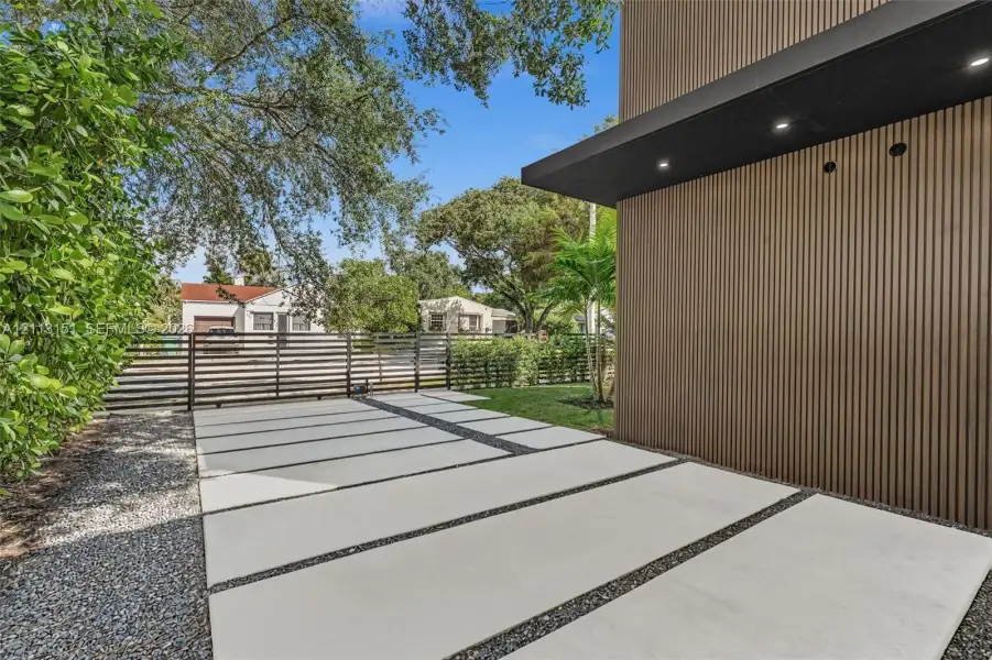 Vertical wood-slat siding, black flat-roof overhang with recessed lighting, and concrete paver driveway with decorative aggregate infill Vertical wood-slat siding, black flat-roof overhang with recessed lighting, and concrete paver driveway with decorative aggregate infill