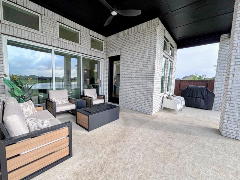 The spacious covered patio features modern outdoor seating, a ceiling fan with large windows offering scenic views of a nearby lake. Perfect for relaxing and entertaining.