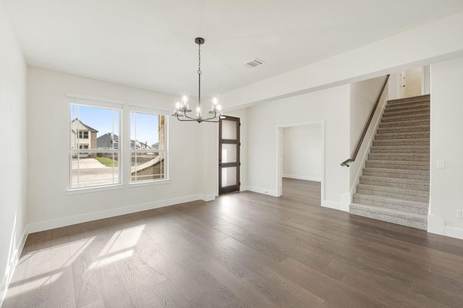 Open living and dining area with hardwood floors, chandelier, dark wood door, and carpeted staircase nearby.