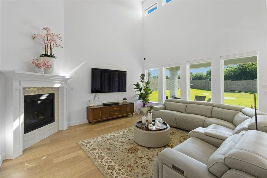 Two-story living area featuring a corner fireplace with white mantle, light wood-finish flooring, and multiple floor-to-ceiling windows overlooking the backyard