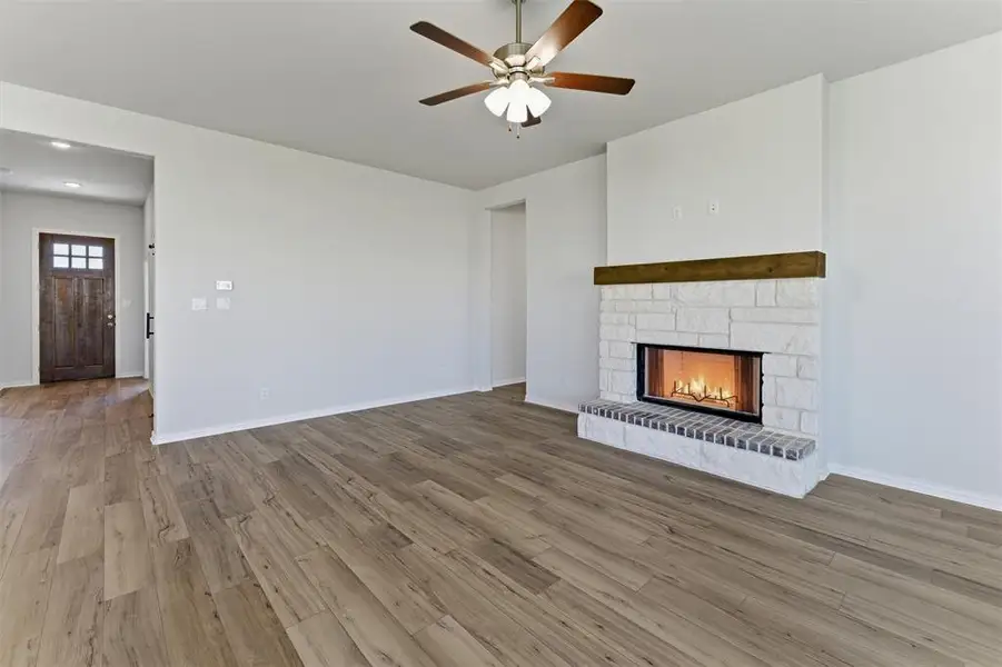 Spacious living area featuring wood-finish flooring, a white stone fireplace with a dark wood mantel, and a ceiling fan with integrated lighting