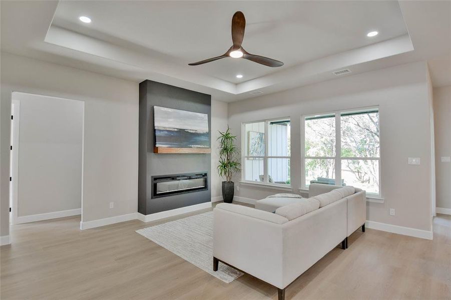 Living room featuring a tray ceiling, a glass covered fireplace, ceiling fan, light wood finished floors, and recessed lighting Living room featuring a tray ceiling, a glass covered fireplace, ceiling fan, light wood finished floors, and recessed lighting