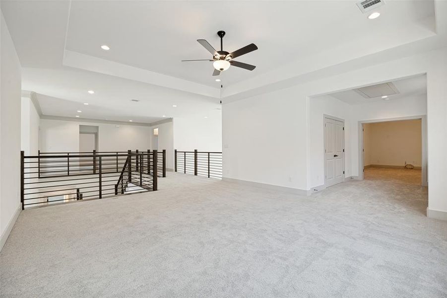 Expansive carpeted loft with a modern horizontal metal railing, tray ceiling, recessed lighting, and a ceiling fan Expansive carpeted loft with a modern horizontal metal railing, tray ceiling, recessed lighting, and a ceiling fan