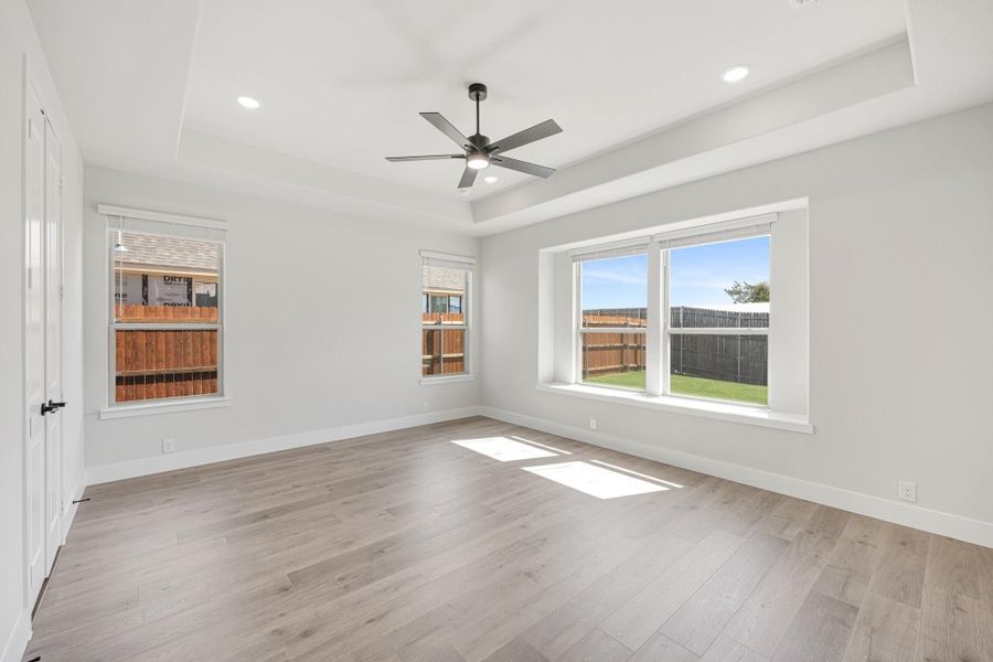 Empty bedroom with tray ceiling, ceiling fan, hardwood floors, and three windows with backyard view Empty bedroom with tray ceiling, ceiling fan, hardwood floors, and three windows with backyard view