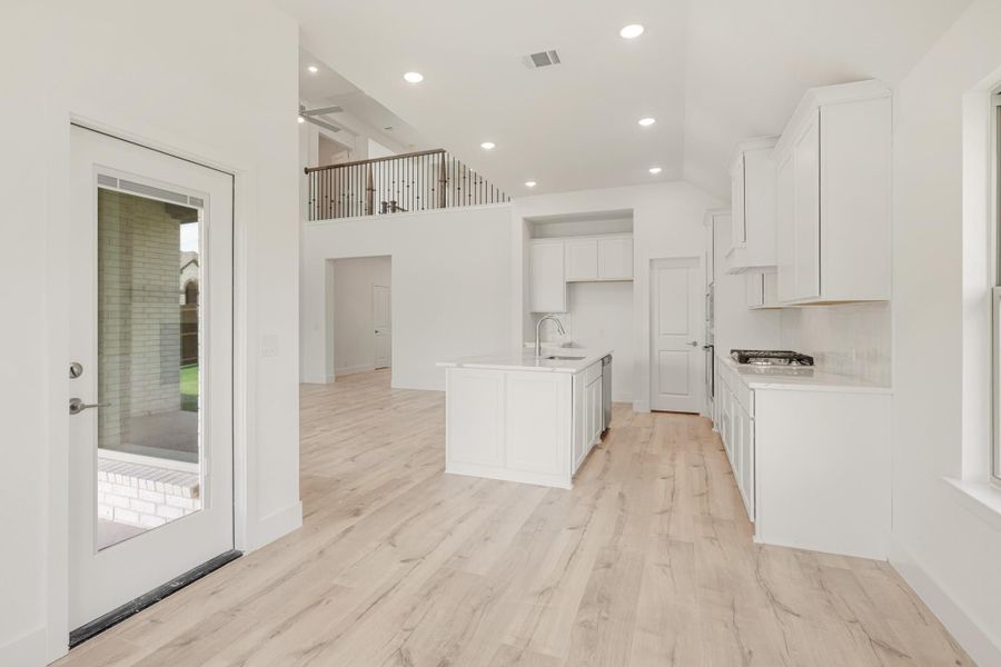 Open kitchen with white cabinets, center island, and light wood floors, open to living area with second-floor balcony visible