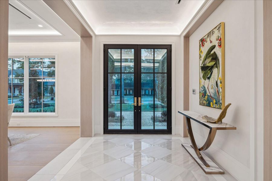 An elegant entry foyer anchored by a steel and glass double doors, polished large-format checkered flooring, polished plaster ceiling with LED lighting and White-Oak Rift Sawn details, setting a bright, sophisticated, and welcoming first impression.