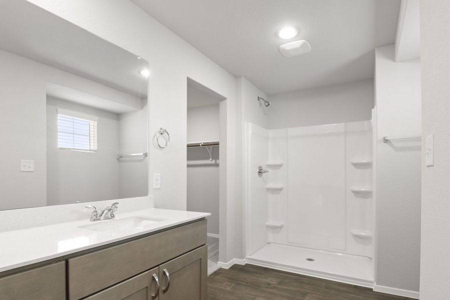 Image of a primary bathroom with a white vinyl walk-in shower, a connecting walk-in closet, and a white vanity with a large mirror