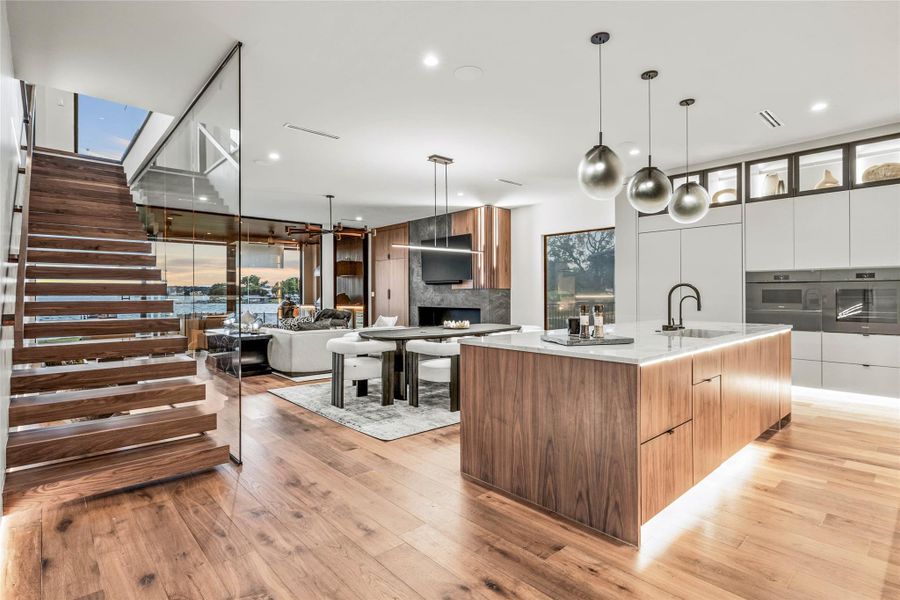 Open-concept interior featuring wood-finish flooring, a floating staircase with a glass railing, a kitchen island with integrated lighting, built-in kitchen appliances, and recessed lighting throughout