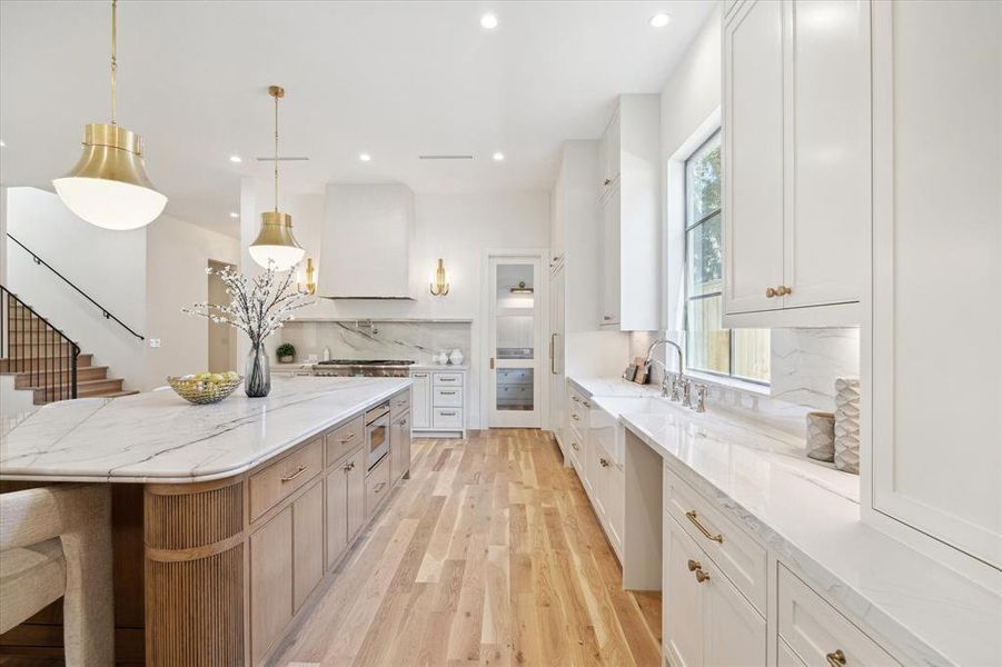 The kitchen island with no sink showcases a double bullnose countertop, white oak details, plenty of storage and brass pendants. The kitchen vent-hood is completed with a plaster finish and brass sconces on each side.