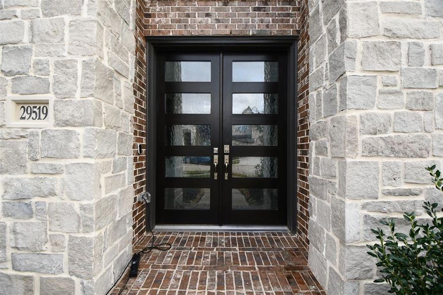 This photo showcases a modern front entry with a dark double door featuring glass panels, set against a robust stone and brick facade. The house number is prominently displayed on the left, offering a welcoming and stylish entrance.