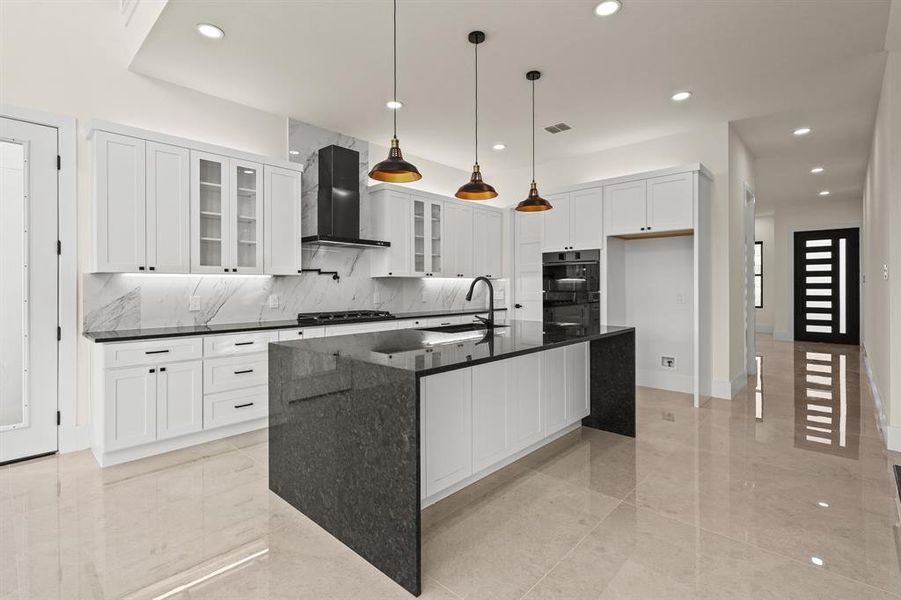 Kitchen featuring wall chimney range hood, sink, a kitchen island with sink, white cabinetry, and dark stone countertops