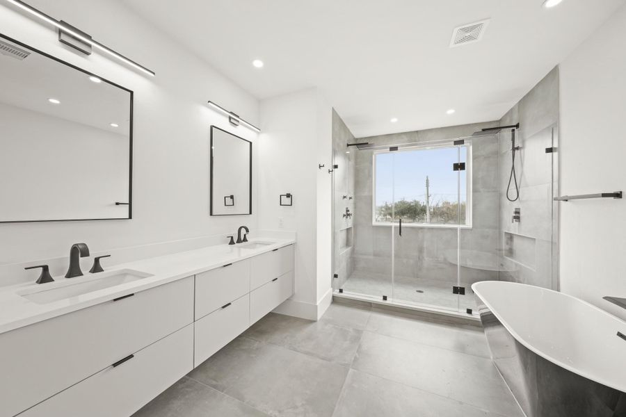 Beautifully appointed, the owner's private bath includes a floating vanity supporting a Quartz countertop, dual under mount sinks, oversized black framed mirrors with lighting above with tile floors.Full view of the primary bathroom showcasing large tile floors, dual sinks with a quartz counter, black fixtures, black-framed mirrors with decorative lighting, a freestanding tub with a waterfall faucet, and an 8-foot glass-enclosed shower with recessed lights and a fixed window.