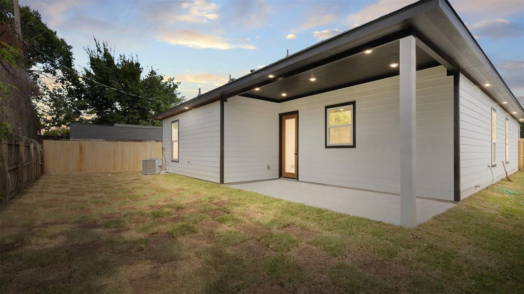 This photo showcases a modern home's backyard with a clear lawn, wooden fencing, and a covered patio area with recessed lighting. The exterior features clean lines and a neutral color palette, offering a private and inviting outdoor space.