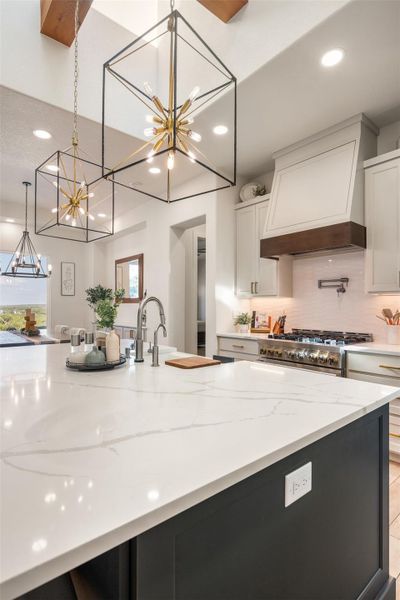 Kitchen with white cabinets, custom exhaust hood, hanging light fixtures, stainless steel range, and light stone counters Kitchen with white cabinets, custom exhaust hood, hanging light fixtures, stainless steel range, and light stone counters