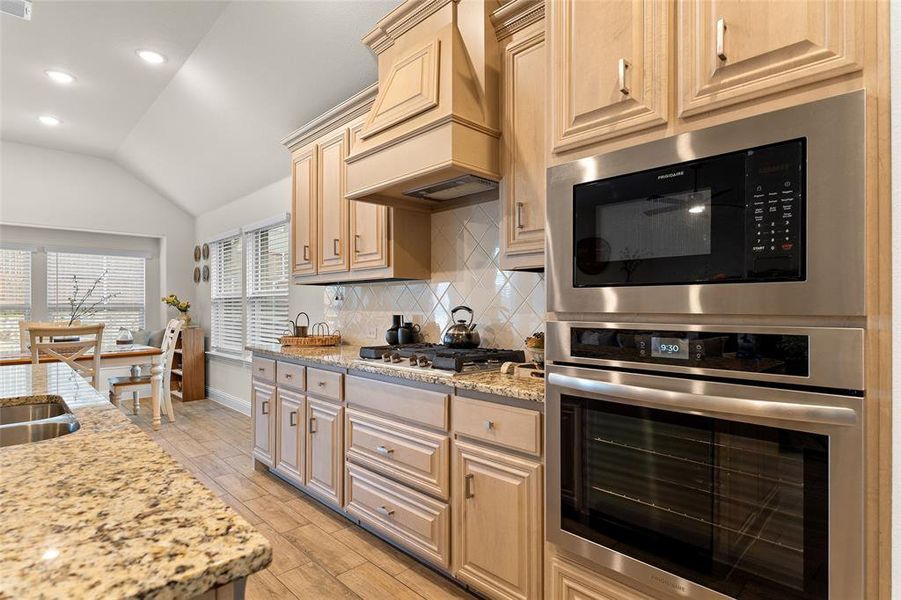 Kitchen with stainless steel appliances, light stone countertops, vaulted ceiling, backsplash, and premium range hood Kitchen with stainless steel appliances, light stone countertops, vaulted ceiling, backsplash, and premium range hood