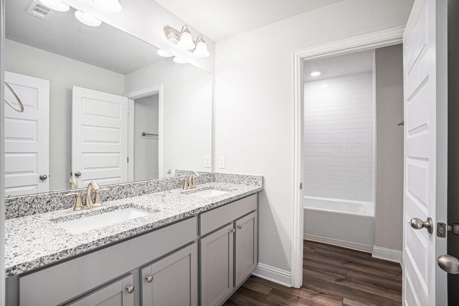 This bathroom features a dual-sink vanity with granite countertops, modern fixtures, and ample cabinet space. It has a large mirror and leads to a separate area with a bathtub and shower, set against a backdrop of clean, white tiles. The luxury vinyl plank flooring adds warmth to the space.