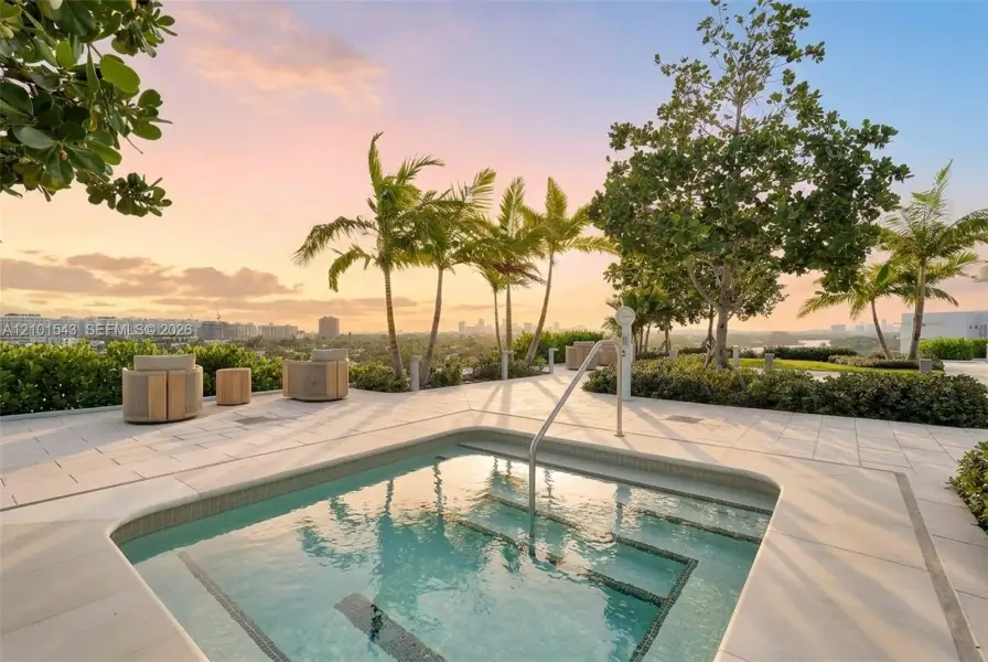 In-ground spa with stepped entry and metal handrail, surrounded by light-toned paver patio, lush perimeter landscaping, and mature palm trees