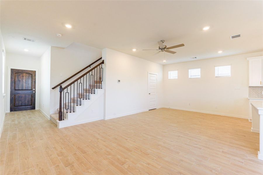 This spacious living area features light wood flooring, a ceiling fan, and ample natural light from several windows. It includes a modern staircase with dark wood accents, recessed lighting, and a view into the kitchen area, creating an open and inviting atmosphere.