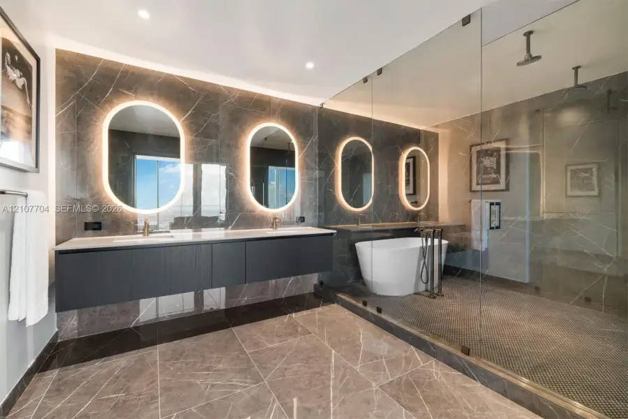 Bathroom featuring a floating double vanity with a light-toned countertop, four wall-mounted illuminated oval mirrors, a freestanding soaking tub, and a large glass-enclosed walk-in shower with stone tile flooring and wall surfacing