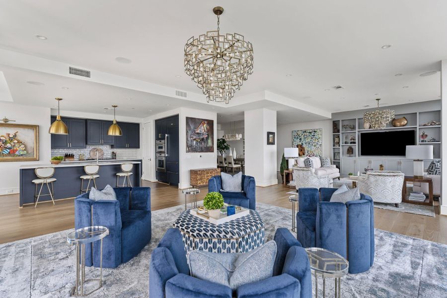 This spacious, open-concept living area features elegant design with a modern kitchen, complete with sleek navy cabinetry and gold accents. The room is highlighted by luxurious lighting fixtures, plush blue seating, and a cozy entertainment space with built-in shelving. Large windows provide abundant natural light, creating a warm and inviting atmosphere. This spacious, open-concept living area features elegant design with a modern kitchen, complete with sleek navy cabinetry and gold accents. The room is highlighted by luxurious lighting fixtures, plush blue seating, and a cozy entertainment space with built-in shelving. Large windows provide abundant natural light, creating a warm and inviting atmosphere.