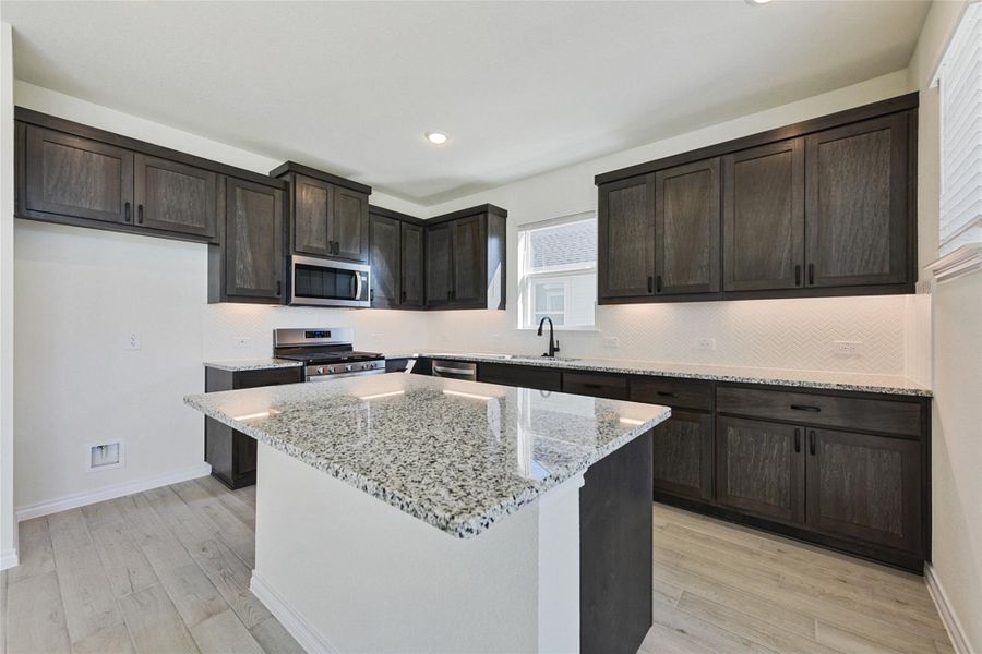 Kitchen featuring tasteful backsplash, appliances with stainless steel finishes, dark brown cabinets, light wood finished floors, and light stone countertops Kitchen featuring tasteful backsplash, appliances with stainless steel finishes, dark brown cabinets, light wood finished floors, and light stone countertops