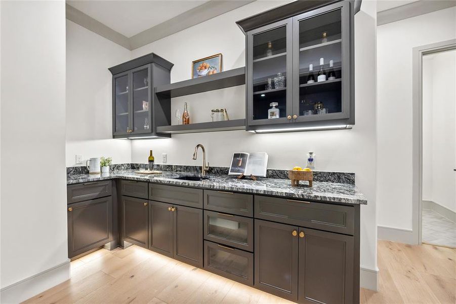 Indoor wet bar with glass fronted cabinets, dark stone counters, open shelves, and light wood finished floors Indoor wet bar with glass fronted cabinets, dark stone counters, open shelves, and light wood finished floors