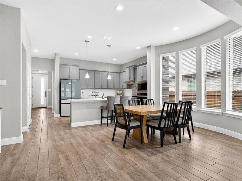 Open-concept living area featuring wood-finish flooring, a kitchen with grey cabinetry and an island with pendant lighting, and a dining space with curved walls and multiple windows Open-concept living area featuring wood-finish flooring, a kitchen with grey cabinetry and an island with pendant lighting, and a dining space with curved walls and multiple windows