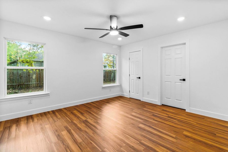 Bright room with hardwood floors, two large windows providing natural light, white walls, a ceiling fan, and two doors, likely leading to closets. Ideal for a bedroom or office.