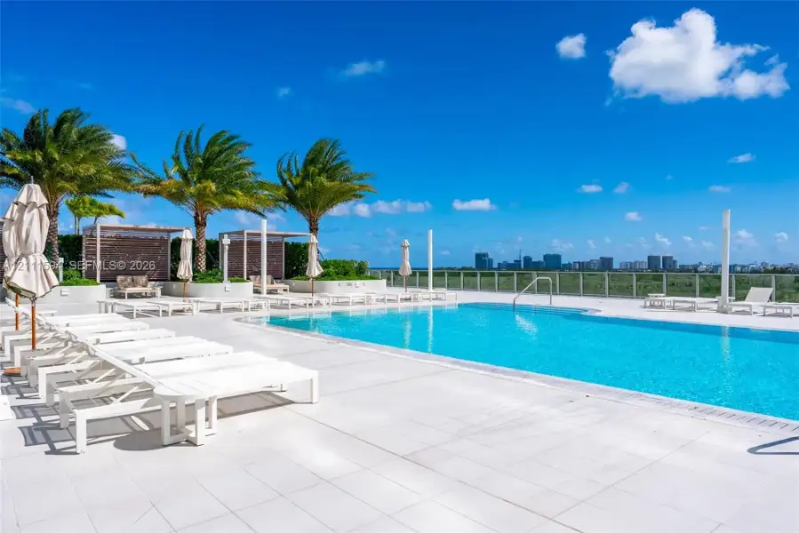 Rooftop pool deck featuring stone tile flooring, perimeter glass railings, built-in cabanas, and mature palm trees Rooftop pool deck featuring stone tile flooring, perimeter glass railings, built-in cabanas, and mature palm trees