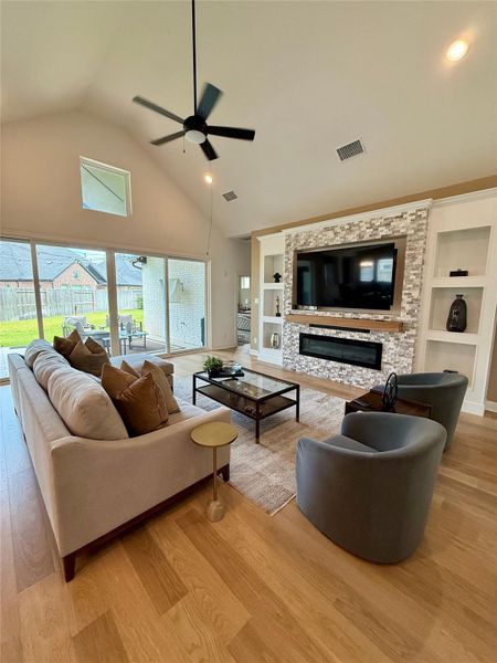 Spacious living room with high ceilings, elegant wood flooring, and custom designed modern stone accent wall featuring a built-in fireplace and TV. Large windows provide ample natural light and offer views of the backyard.