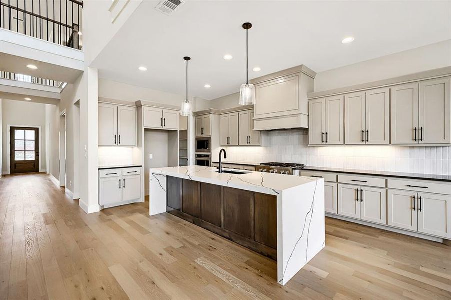 Kitchen featuring light stone counters, pendant lighting, an island with sink, light wood-style floors, and backsplash Kitchen featuring light stone counters, pendant lighting, an island with sink, light wood-style floors, and backsplash