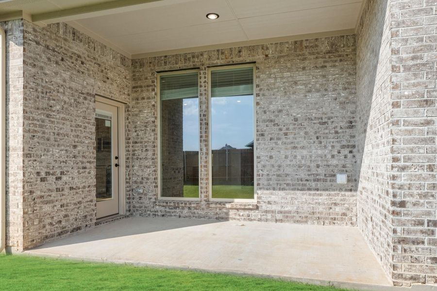 Covered back patio with concrete floor, brick exterior walls, back door, and large windows overlooking a fenced yard.