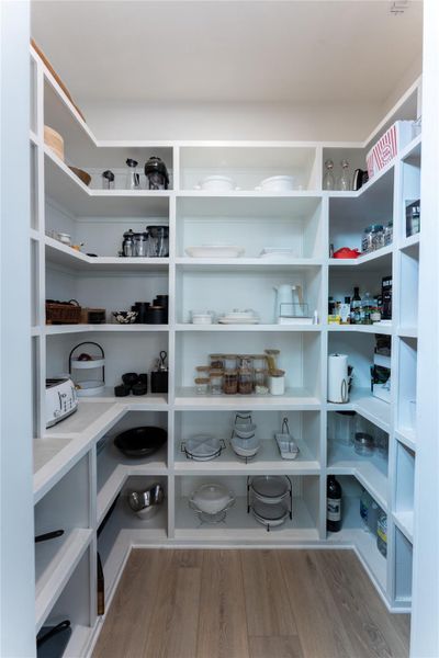 Spacious walk-in pantry featuring extensive white built-in shelving, wrap-around countertop space, and wood-finish flooring Spacious walk-in pantry featuring extensive white built-in shelving, wrap-around countertop space, and wood-finish flooring