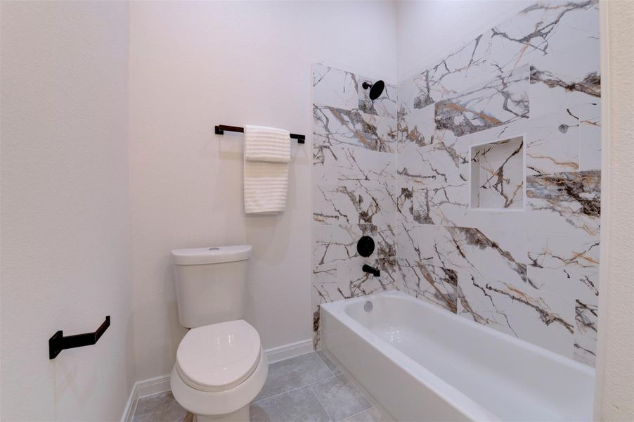 Tub/shower combo with dramatic marble-look tile surround, built-in niche, matte black shower fixtures, and tile flooring.