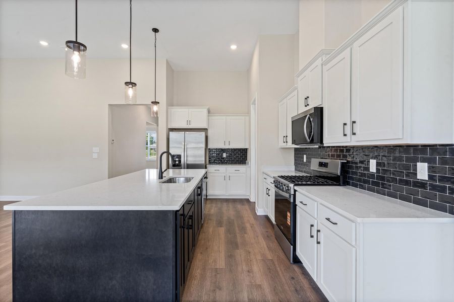 Modern kitchen featuring a large island with sink, stainless steel appliances, white cabinetry, a black subway tile backsplash, and wood-finish flooring Modern kitchen featuring a large island with sink, stainless steel appliances, white cabinetry, a black subway tile backsplash, and wood-finish flooring