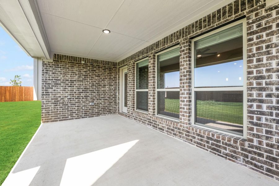 Covered back patio with concrete floor, brick exterior walls, and large windows overlooking a fenced backyard with green lawn. Covered back patio with concrete floor, brick exterior walls, and large windows overlooking a fenced backyard with green lawn.