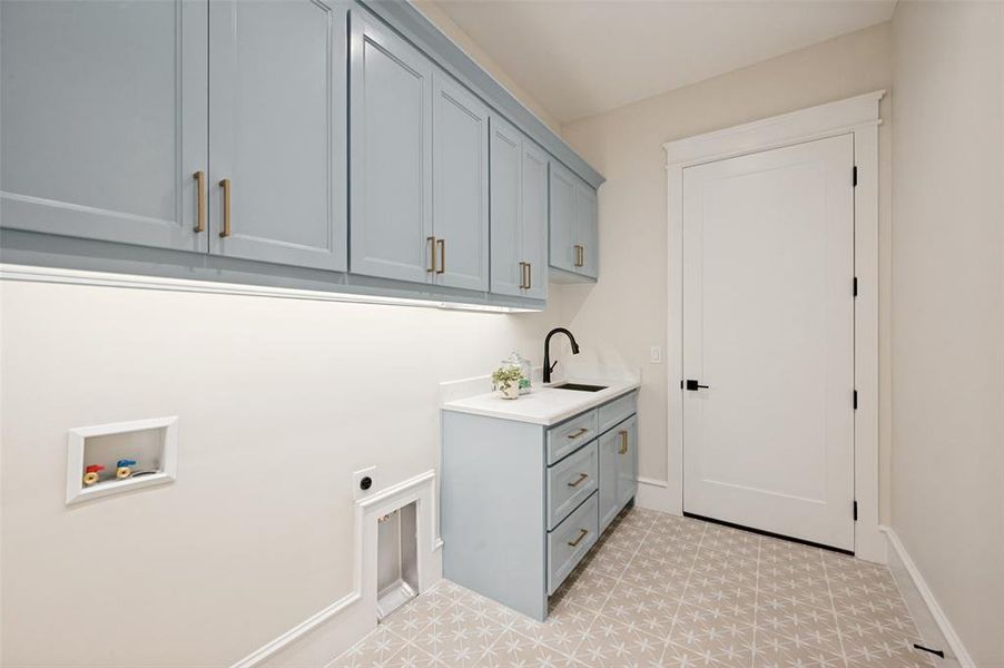 Laundry room featuring light blue cabinetry with gold-finish hardware, a built-in sink with a matte black faucet, and patterned floor tiles